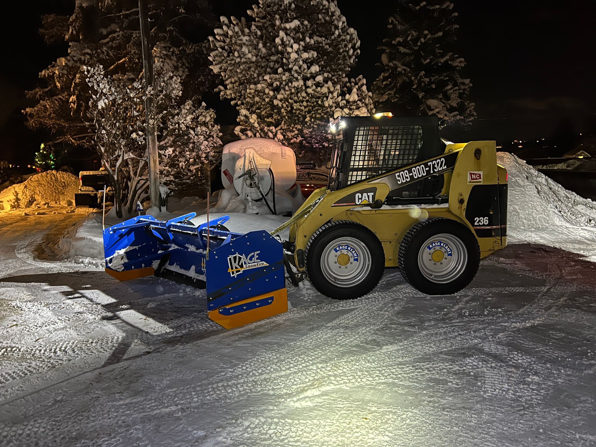A yellow and blue snow plow is parked in the snow at night.
