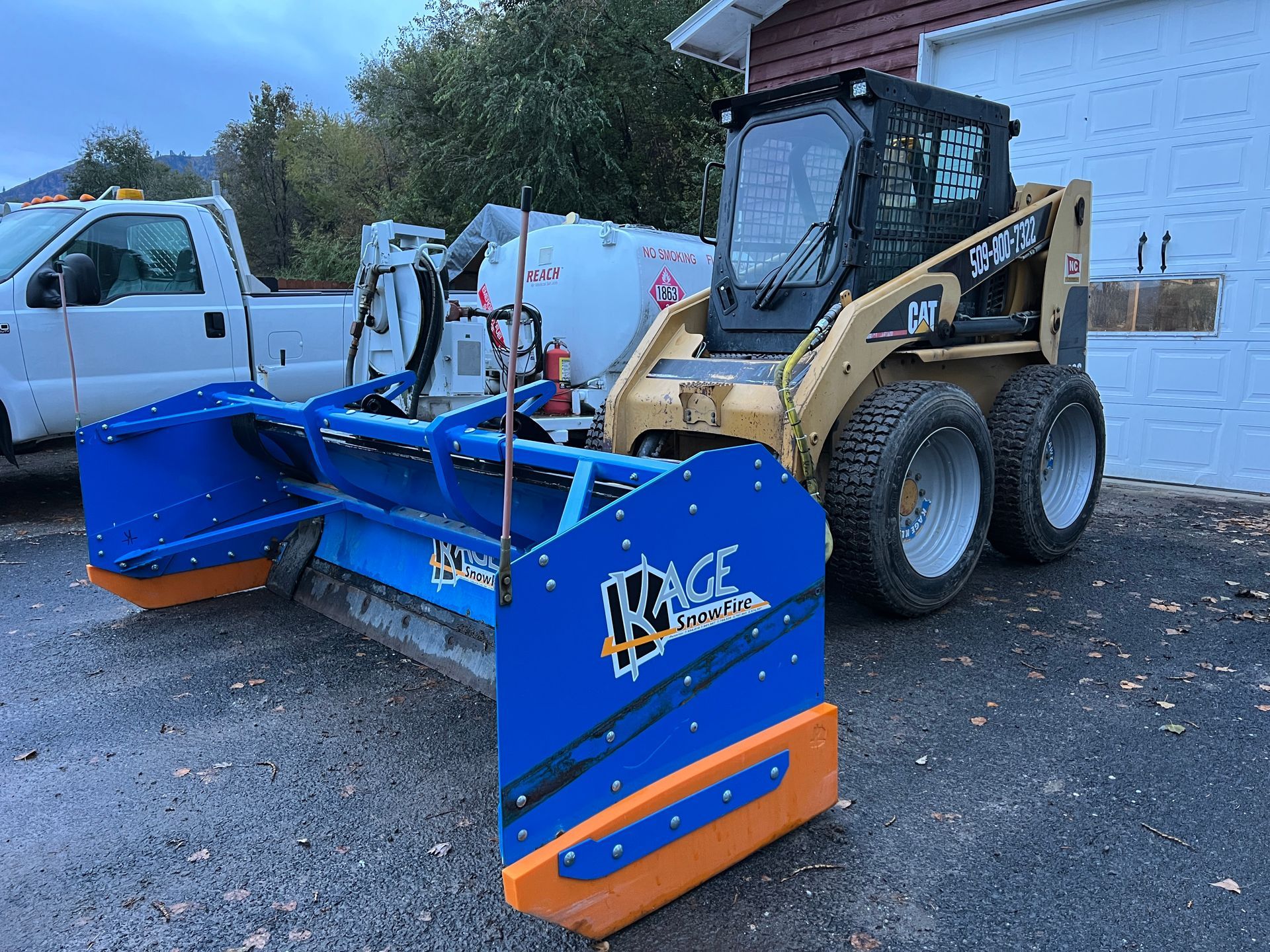 A snow plow is parked in front of a garage next to a truck.