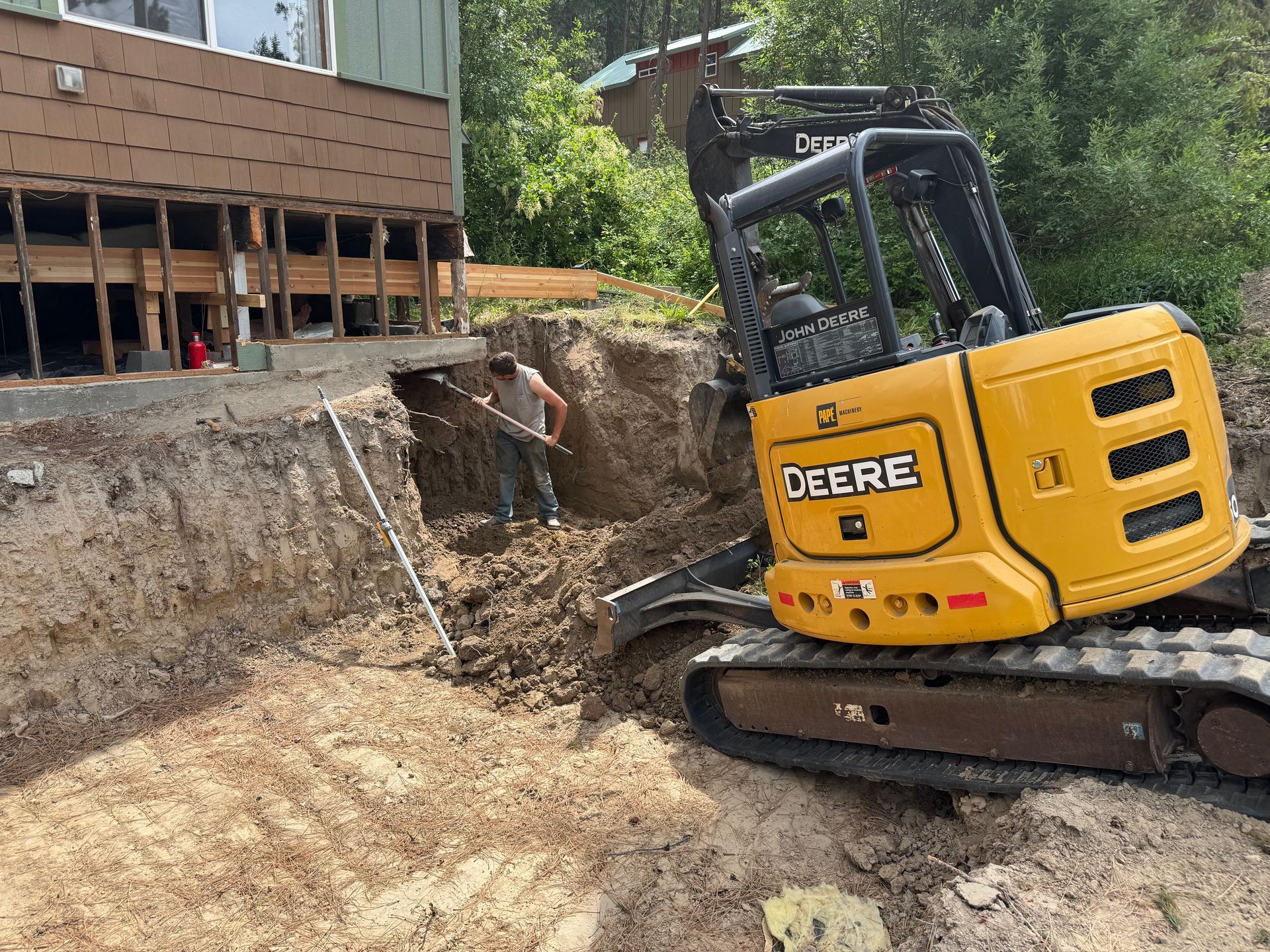 A yellow deere excavator is digging a hole in front of a house.